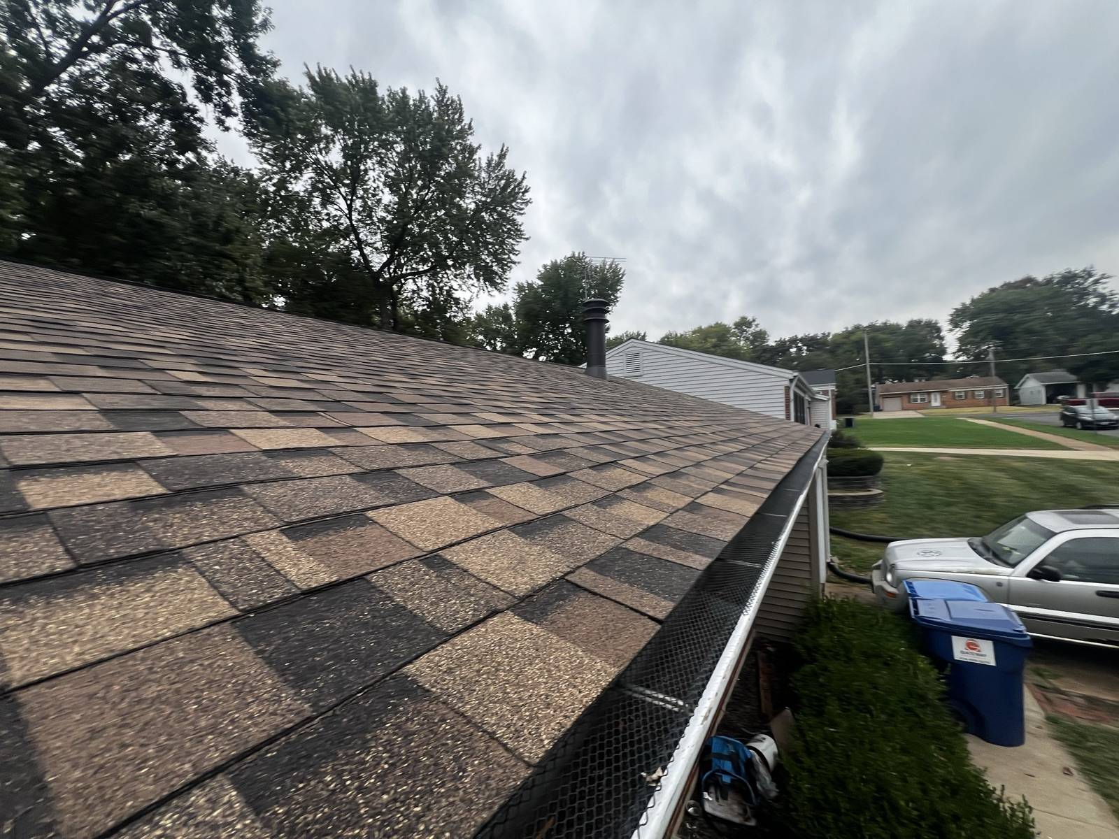 A continuous white gutter run with a black mesh guard along the full eave of a weathered shingle roof, with the driveway and the street below