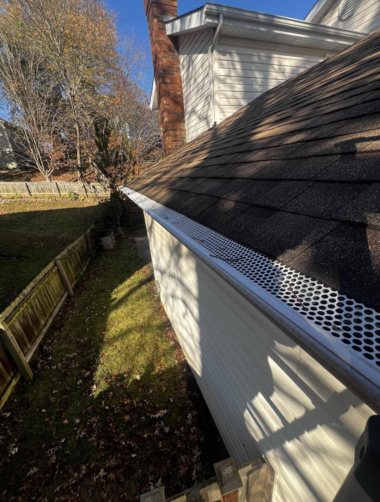 A white perforated metal gutter guard fitted the length of a gutter along the eave of a weathered dark shingle roof, with two bare twigs and a few small dark scraps of debris lying on top of the perforations, a white sided wall and a brick chimney above and a wooden fence and fallen autumn leaves on the lawn below