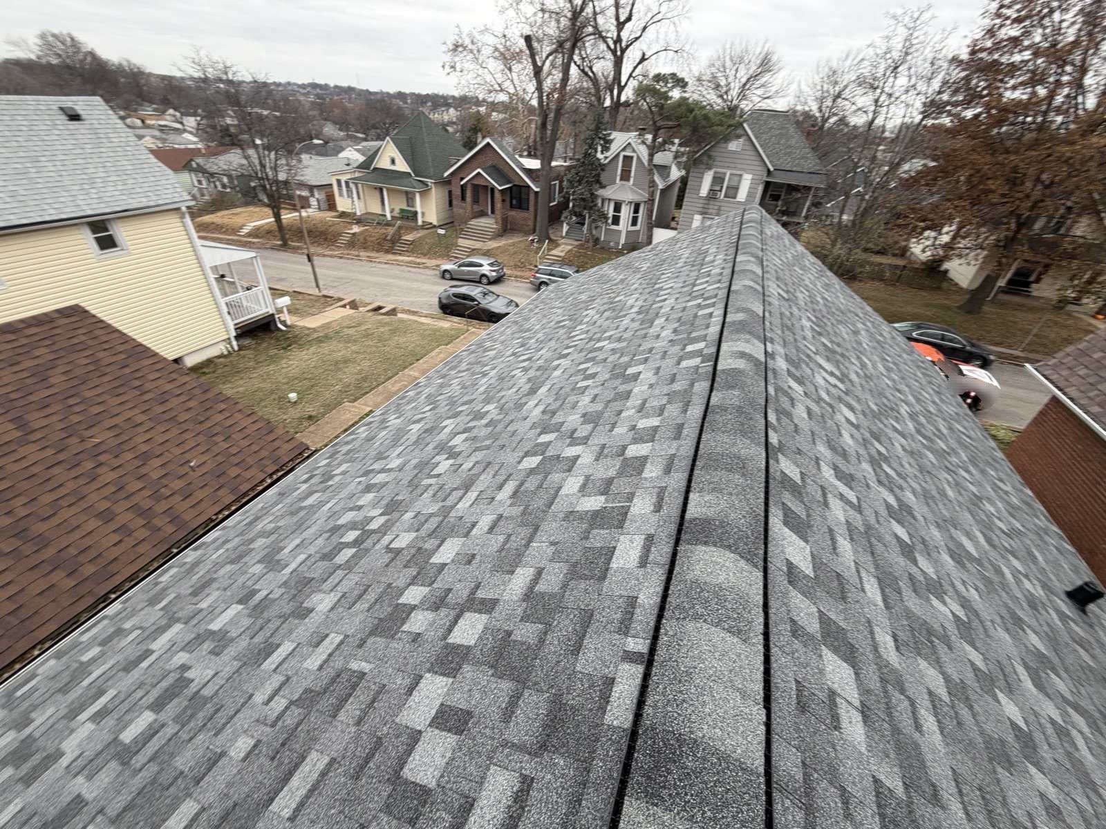 Finished grey architectural shingle roof photographed along the ridge, with a ridge vent running the length and an older St. Louis street below